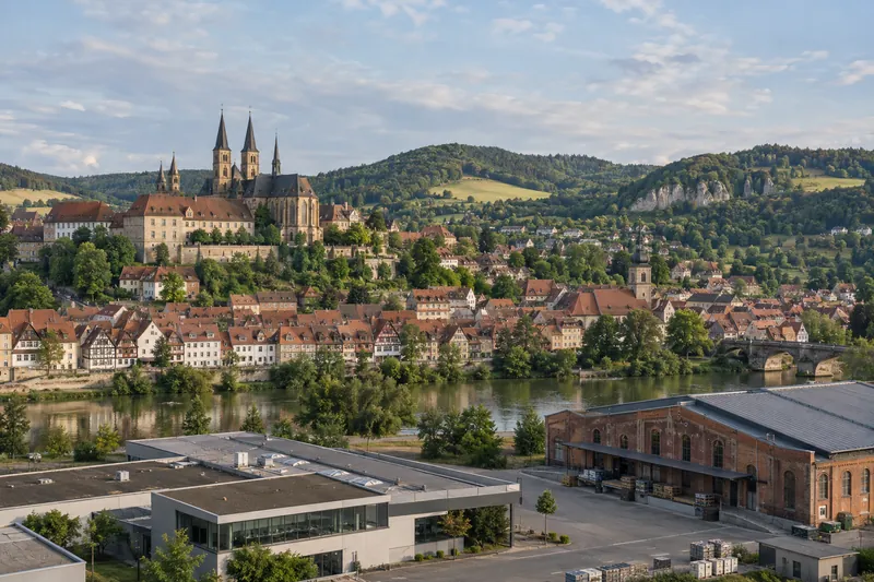 Fränkische Kleinstadt mit Fachwerk und Sandstein, davor Gewerbebau und Backsteinhalle, dahinter Hügel und Domtürme