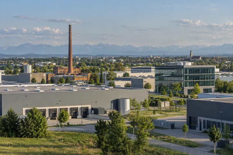 Gewerbegebiet am Stadtrand von Augsburg mit Lagerhallen, Bürogebäude und altem Fabrikschornstein, Alpenvorland im Hintergrund