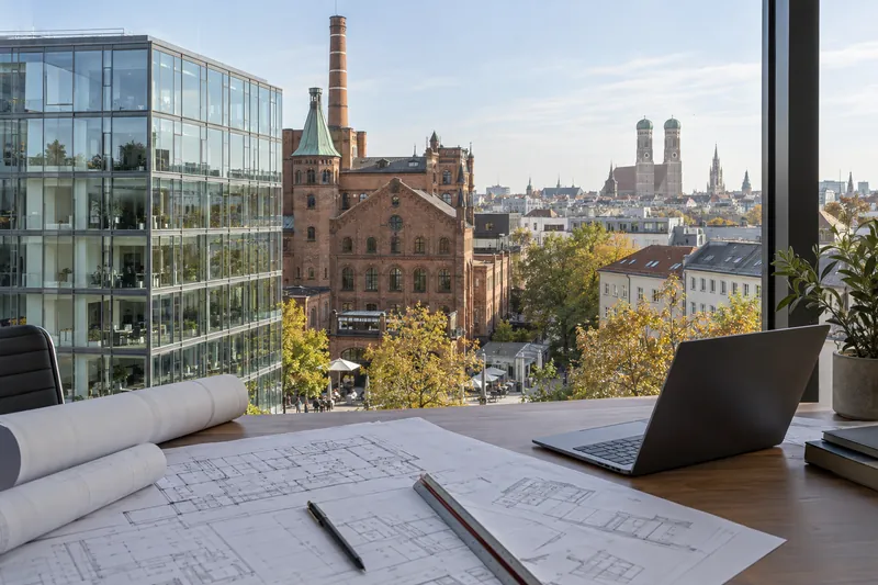 Moderner Büroneubau neben einem Backstein-Brauereigebäude, im Hintergrund die Silhouette Münchens, im Vordergrund Pläne auf einem Tisch