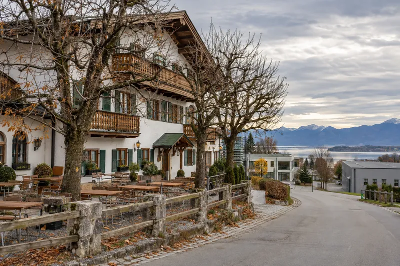 Oberbayerischer Gasthof mit Biergarten, dahinter Gewerbebauten und Alpenvorland am Horizont