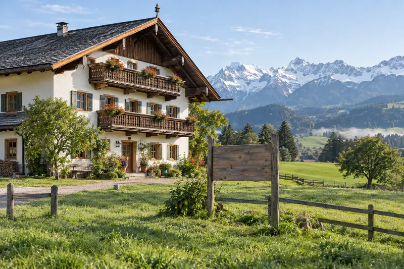 Oberbayerischer Gasthof mit Holzbalkonen vor Alpenkulisse, davor eine Wiese mit leerer Holztafel