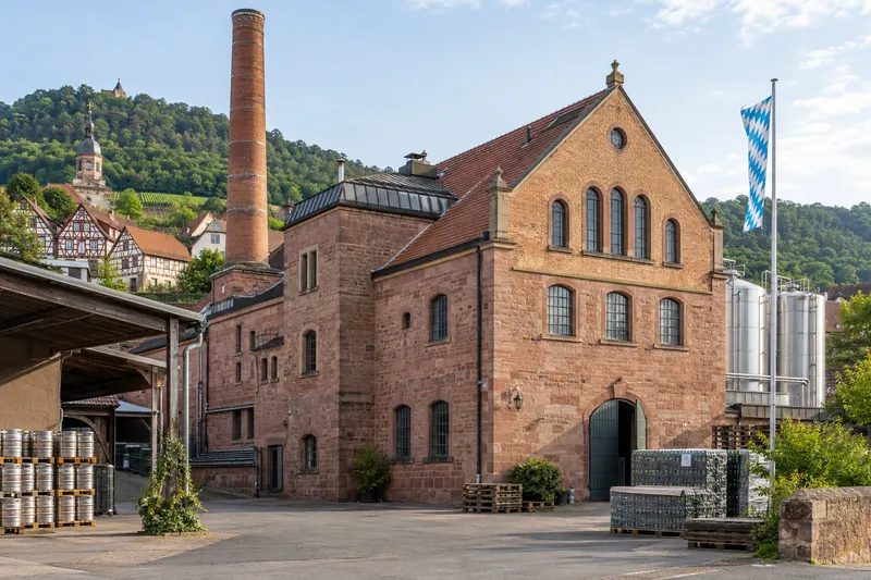 Fränkische Brauerei aus Sandstein und Backstein mit hohem Schornstein, im Hintergrund Hügel und Fachwerkhäuser
