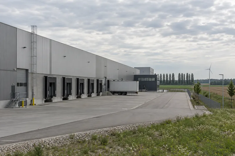 Große Produktionshalle mit Metallfassade und Verladerampe in einem Gewerbegebiet bei Ingolstadt, Pappelreihe und Windrad im Hintergrund