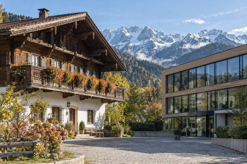 Oberbayerischer Gasthof mit Holzbalkon neben einem modernen Bürogebäude, im Hintergrund die Alpen
