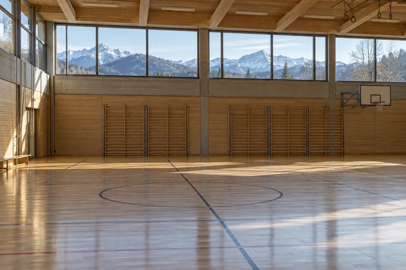 Leere Sporthalle mit Holzboden und hohen Fensterbändern, durch die Alpenvorland-Hügel zu sehen sind