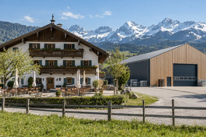 Bayerischer Gasthof mit Biergarten neben einer modernen Gewerbehalle vor Alpenkulisse