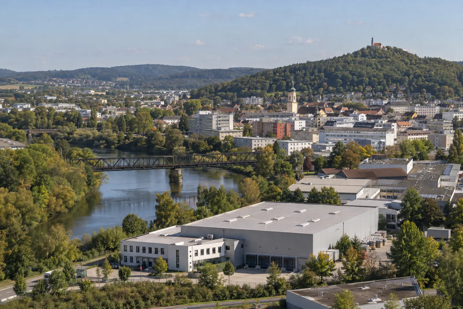 Gewerbehalle mit Bürotrakt in Schwandorf, dahinter die Naab mit Bahnbrücke und der bewaldete Kreuzberg