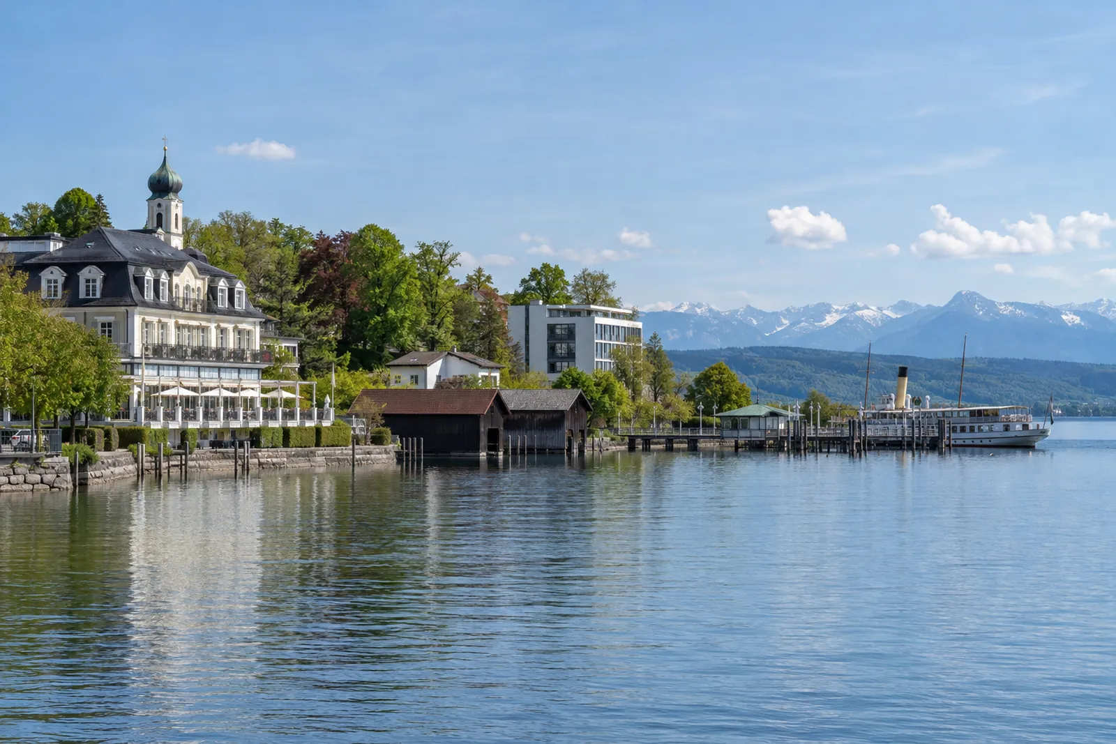 Seepromenade in Starnberg mit Hotel, Bootshäusern und Blick über den Starnberger See Richtung Alpen